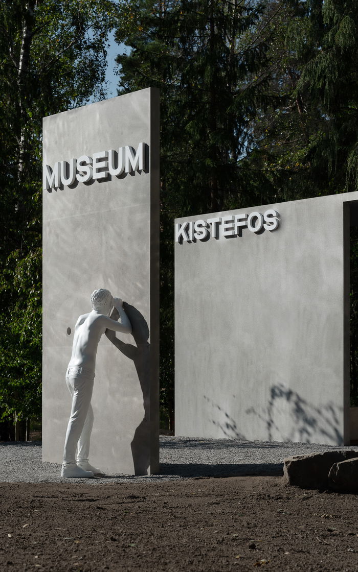 Skulpturen viser en hvit mann som kikker gjennom et hull i veggen på inngangspartiet til Kistefos.